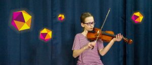 Darcy, a young person, sitting an dplaying violin infront of dark curtain, with colourful dodecahedrons floating around them